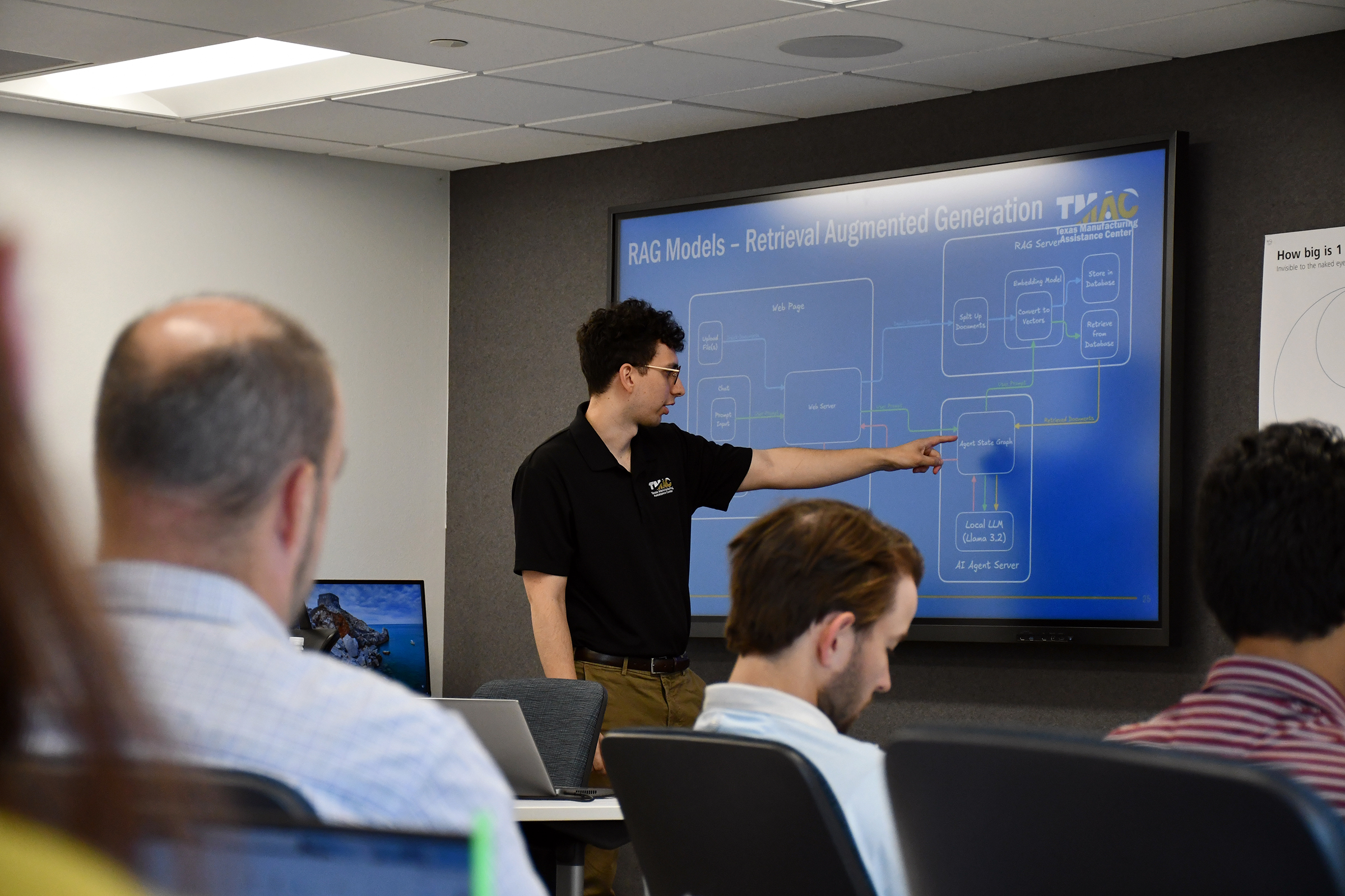 A young male student in a black shirt leading a demonstration, pointing to a presentation screen.