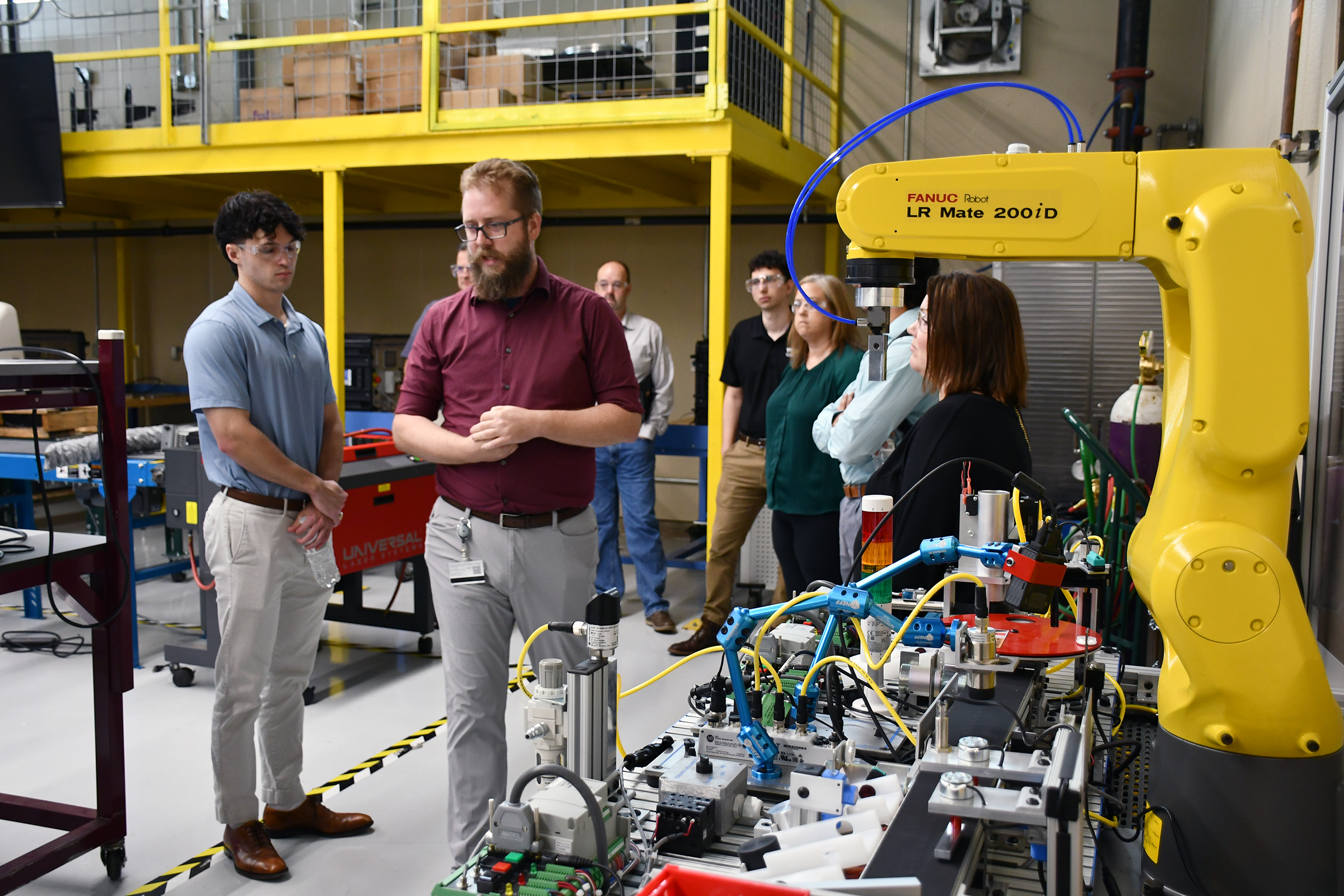 Organizers, instructors, and students discussing a large yellow robotic arm and experimentation setup.